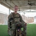 A military service member in full MultiCam/OCP camouflage uniform and tactical plate carrier vest poses with a military working dog (German Shepherd) on artificial turf under a covered outdoor area. The handler is kneeling behind the dog, which is in a down position with its tongue out. The setting appears to be a forward operating base or military installation kennel facility.
