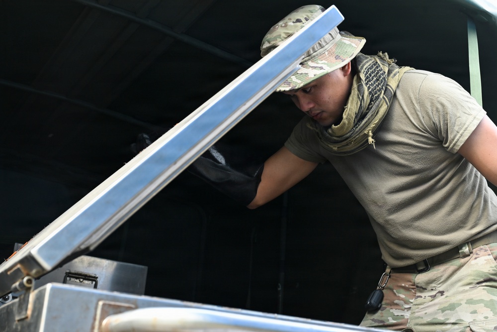 A military personnel member wearing MultiCam/OCP camouflage uniform, boonie hat, and shemagh scarf is working with a piece of metal equipment, appearing to lift or adjust a metal panel or lid. The individual is wearing black tactical gloves and is focused on the task at hand. The setting appears to be a field or operational environment with a dark canopy or tent structure visible in the background.