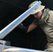 A military personnel member wearing MultiCam/OCP camouflage uniform, boonie hat, and shemagh scarf is working with a piece of metal equipment, appearing to lift or adjust a metal panel or lid. The individual is wearing black tactical gloves and is focused on the task at hand. The setting appears to be a field or operational environment with a dark canopy or tent structure visible in the background.