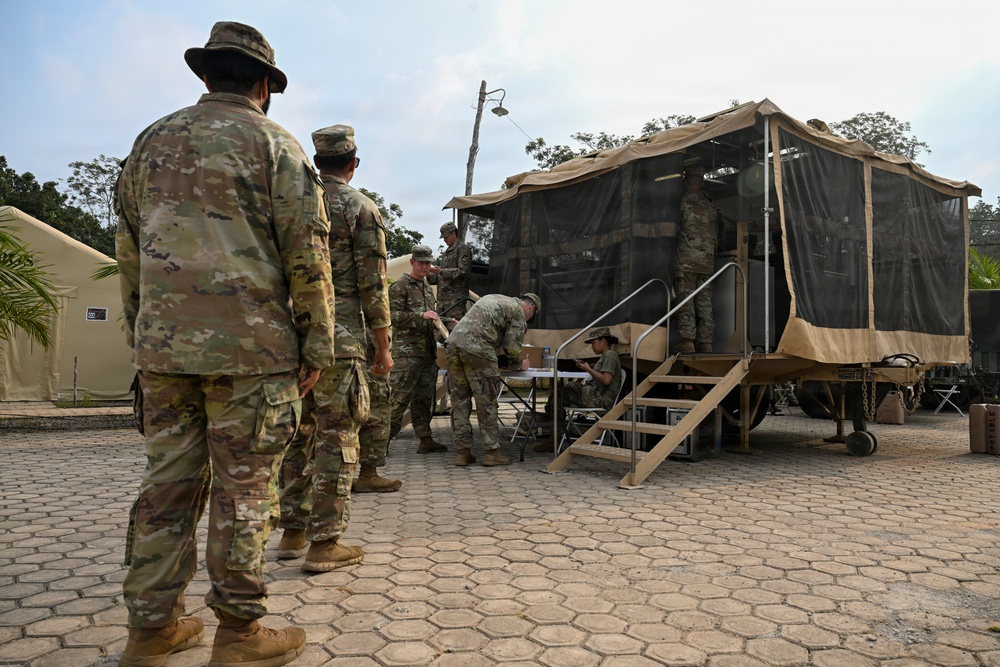 Military personnel in OCP (Operational Camouflage Pattern) uniforms are gathered at what appears to be a forward operating base or field camp. Soldiers are queued at a field processing or administrative station set up near a mobile military shelter unit. The setting includes tactical tents, a paved hexagonal-tile courtyard, and tropical vegetation in the background.