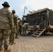 Military personnel in OCP (Operational Camouflage Pattern) uniforms are gathered at what appears to be a forward operating base or field camp. Soldiers are queued at a field processing or administrative station set up near a mobile military shelter unit. The setting includes tactical tents, a paved hexagonal-tile courtyard, and tropical vegetation in the background.