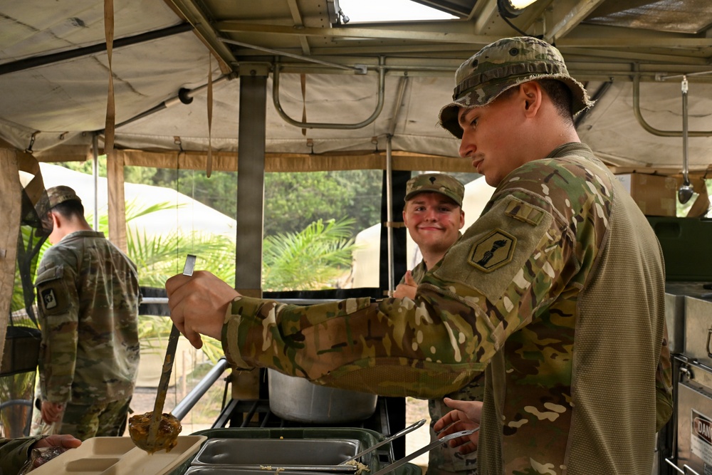 Military personnel are operating a field kitchen under a canvas tent structure in what appears to be a tropical or subtropical environment. A soldier in the foreground is serving food using a ladle into metal serving trays, while a second soldier smiles in the background. A third soldier is visible from behind on the left side of the frame.