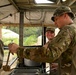 Military personnel are operating a field kitchen under a canvas tent structure in what appears to be a tropical or subtropical environment. A soldier in the foreground is serving food using a ladle into metal serving trays, while a second soldier smiles in the background. A third soldier is visible from behind on the left side of the frame.