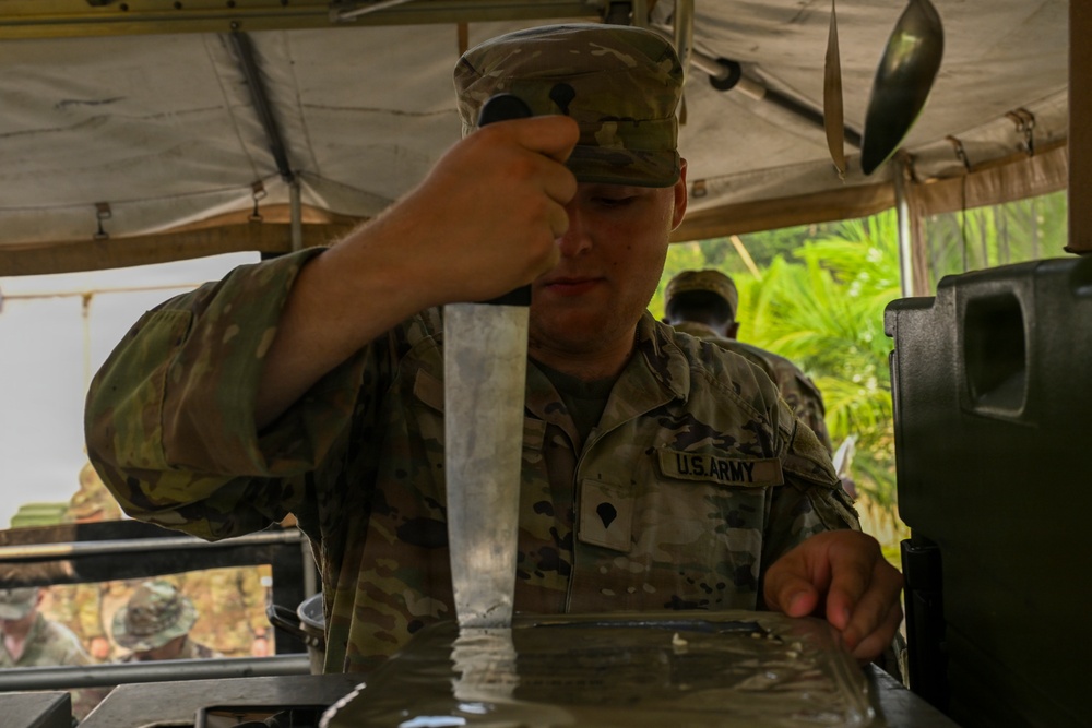 A U.S. Army soldier in MultiCam/OCP camouflage uniform is working at what appears to be a field kitchen or water purification station under a canvas shelter. The soldier is handling a metal implement over a water-filled container or basin. The setting appears to be a tropical or jungle environment based on the vegetation visible in the background.