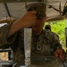 A U.S. Army soldier in MultiCam/OCP camouflage uniform is working at what appears to be a field kitchen or water purification station under a canvas shelter. The soldier is handling a metal implement over a water-filled container or basin. The setting appears to be a tropical or jungle environment based on the vegetation visible in the background.