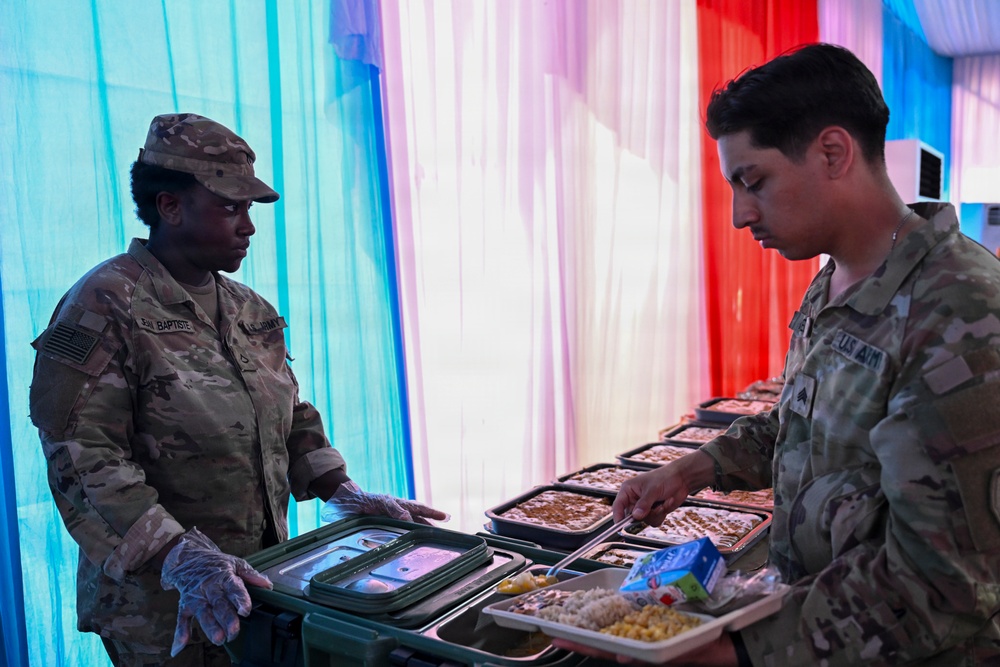 Two U.S. Army soldiers are engaged in a food service operation, likely at a military dining facility or field kitchen. One soldier wearing gloves appears to be serving or distributing meal trays to another soldier. The background features colorful draped fabric in red, white, and blue, suggesting a celebratory or holiday meal event.