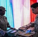 Two U.S. Army soldiers are engaged in a food service operation, likely at a military dining facility or field kitchen. One soldier wearing gloves appears to be serving or distributing meal trays to another soldier. The background features colorful draped fabric in red, white, and blue, suggesting a celebratory or holiday meal event.