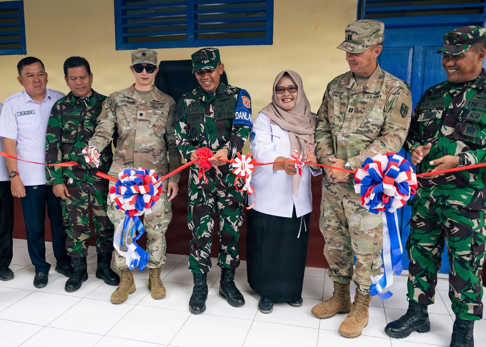 A ribbon-cutting ceremony is taking place at what appears to be a newly completed or renovated building, involving both U.S. Army personnel and Indonesian military (TNI) officers, along with a civilian woman in a hijab. The participants are holding a red ribbon decorated with red, white, and blue bows, suggesting a joint U.S.-Indonesia military cooperation event. Name tapes visible on U.S. Army uniforms include 'GOLDSTEIN' and 'CARRION', and Indonesian uniforms display 'TNI' and 'DANLAN' markings.
