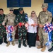 A ribbon-cutting ceremony is taking place at what appears to be a newly completed or renovated building, involving both U.S. Army personnel and Indonesian military (TNI) officers, along with a civilian woman in a hijab. The participants are holding a red ribbon decorated with red, white, and blue bows, suggesting a joint U.S.-Indonesia military cooperation event. Name tapes visible on U.S. Army uniforms include 'GOLDSTEIN' and 'CARRION', and Indonesian uniforms display 'TNI' and 'DANLAN' markings.