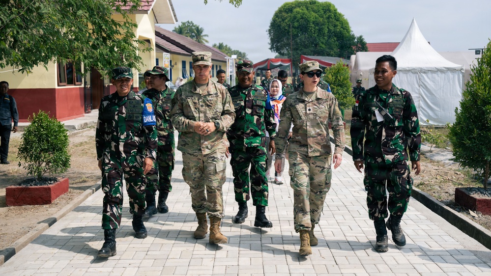 A group of military personnel from at least two different nations are walking together along a paved brick pathway in what appears to be a military compound or base. Two individuals in MultiCam/OCP uniforms consistent with U.S. Army dress are walking alongside several soldiers wearing green camouflage uniforms consistent with Indonesian Armed Forces (TNI) pattern. The setting includes low buildings, landscaped shrubs, and a white event tent in the background.