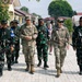 A group of military personnel from at least two different nations are walking together along a paved brick pathway in what appears to be a military compound or base. Two individuals in MultiCam/OCP uniforms consistent with U.S. Army dress are walking alongside several soldiers wearing green camouflage uniforms consistent with Indonesian Armed Forces (TNI) pattern. The setting includes low buildings, landscaped shrubs, and a white event tent in the background.