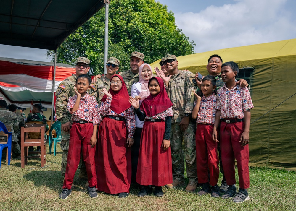 A group of U.S. Army soldiers in MultiCam/OCP camouflage uniforms pose for a photo with local schoolchildren wearing red-and-white uniforms and headscarves, likely during a humanitarian or community engagement mission. The setting appears to be an outdoor event area with military tents and a red-and-white flag visible in the background, consistent with an Indonesian location. The mood is cheerful, with children and soldiers waving and smiling at the camera.