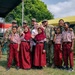 A group of U.S. Army soldiers in MultiCam/OCP camouflage uniforms pose for a photo with local schoolchildren wearing red-and-white uniforms and headscarves, likely during a humanitarian or community engagement mission. The setting appears to be an outdoor event area with military tents and a red-and-white flag visible in the background, consistent with an Indonesian location. The mood is cheerful, with children and soldiers waving and smiling at the camera.