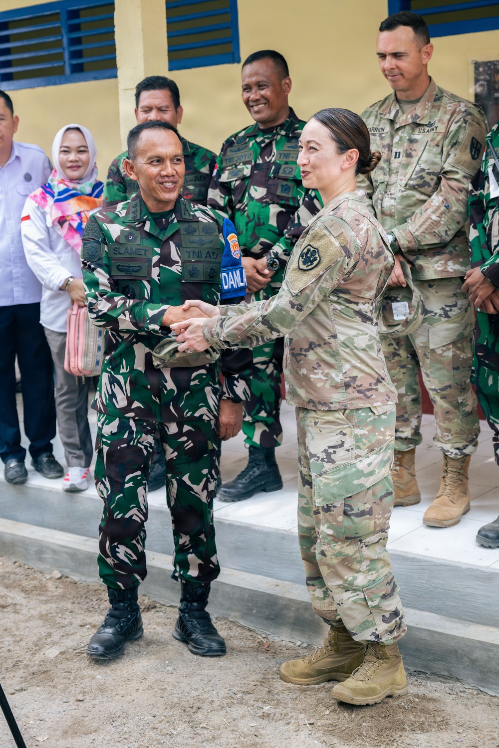 A military handshake ceremony between an Indonesian Army (TNI AD) soldier named 'Slamet' and a U.S. Army female soldier in MultiCam/OCP uniform. The scene appears to be a bilateral military engagement or cooperation event, likely at a newly constructed or renovated building. Additional Indonesian and U.S. military personnel are visible in the background alongside civilian observers.