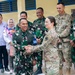 A military handshake ceremony between an Indonesian Army (TNI AD) soldier named 'Slamet' and a U.S. Army female soldier in MultiCam/OCP uniform. The scene appears to be a bilateral military engagement or cooperation event, likely at a newly constructed or renovated building. Additional Indonesian and U.S. military personnel are visible in the background alongside civilian observers.