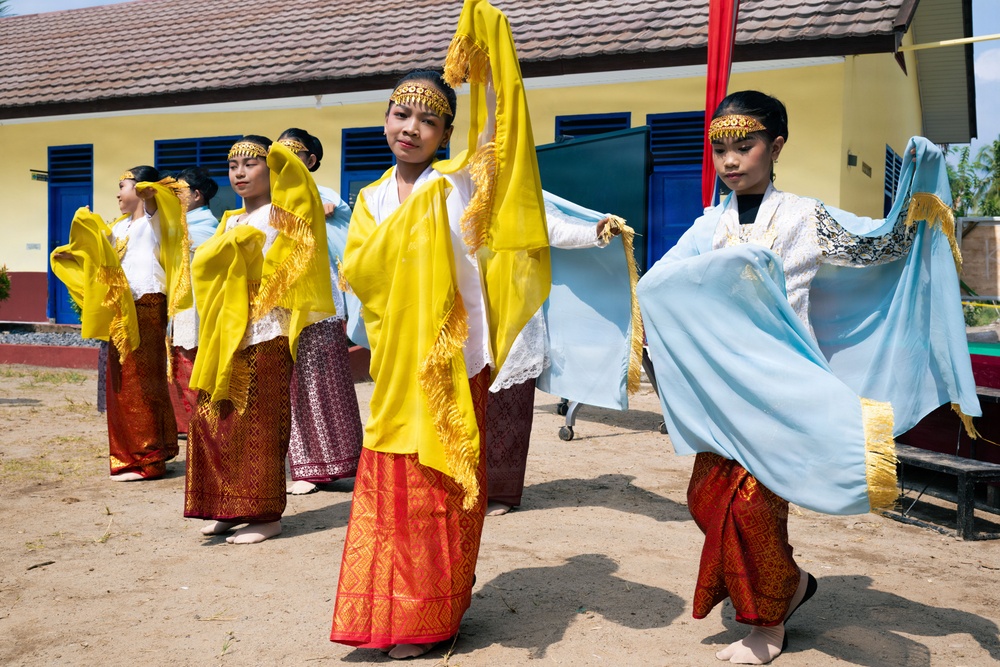 A group of young female dancers are performing a traditional Southeast Asian dance outdoors in front of a yellow building with blue shutters. They are dressed in matching traditional costumes featuring white lace tops, red and gold batik sarongs, gold headbands, and colorful scarves (yellow and light blue) with gold fringe trim. The performance appears to be taking place on a dirt courtyard, likely at a school or community event.
