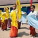 A group of young female dancers are performing a traditional Southeast Asian dance outdoors in front of a yellow building with blue shutters. They are dressed in matching traditional costumes featuring white lace tops, red and gold batik sarongs, gold headbands, and colorful scarves (yellow and light blue) with gold fringe trim. The performance appears to be taking place on a dirt courtyard, likely at a school or community event.