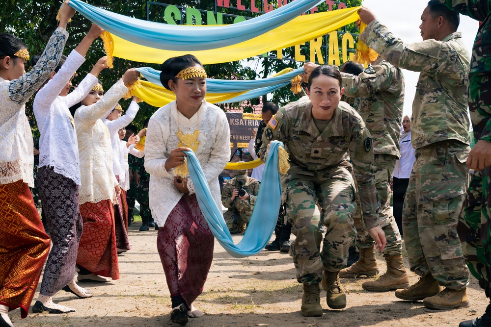 The image depicts a cultural welcome ceremony where US Army soldiers in MultiCam uniforms are participating alongside local women dressed in traditional Indonesian attire (kebaya and batik sarongs). Participants are walking under an arch formed by blue and yellow fabric scarves held aloft by both soldiers and local performers. The event appears to be a military cooperation or humanitarian engagement exercise, as suggested by a partially visible banner referencing 'Engineer' operations.