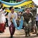 The image depicts a cultural welcome ceremony where US Army soldiers in MultiCam uniforms are participating alongside local women dressed in traditional Indonesian attire (kebaya and batik sarongs). Participants are walking under an arch formed by blue and yellow fabric scarves held aloft by both soldiers and local performers. The event appears to be a military cooperation or humanitarian engagement exercise, as suggested by a partially visible banner referencing 'Engineer' operations.