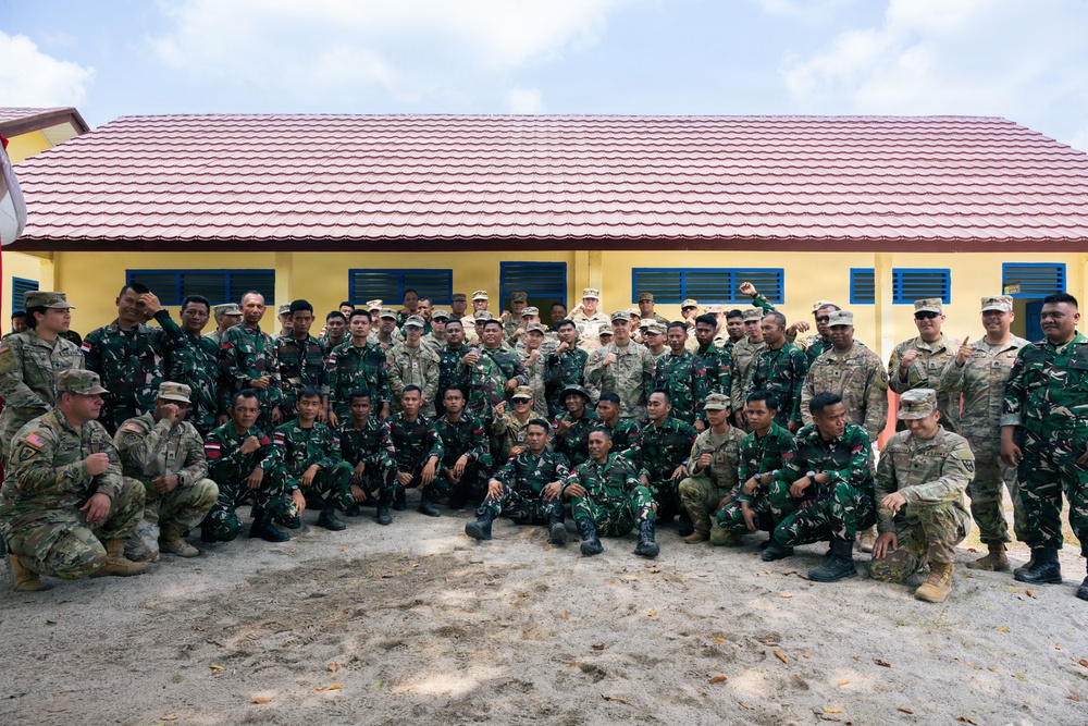 A large group of military personnel from two distinct forces pose together for a formal group photograph in front of a yellow building with a red tile roof. The group includes soldiers in US Army MultiCam/OCP camouflage uniforms and soldiers in green woodland camouflage uniforms consistent with Indonesian Armed Forces (TNI) attire, some wearing Indonesian flag patches. This appears to be a joint military exercise or bilateral training event.