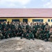 A large group of military personnel from two distinct forces pose together for a formal group photograph in front of a yellow building with a red tile roof. The group includes soldiers in US Army MultiCam/OCP camouflage uniforms and soldiers in green woodland camouflage uniforms consistent with Indonesian Armed Forces (TNI) attire, some wearing Indonesian flag patches. This appears to be a joint military exercise or bilateral training event.