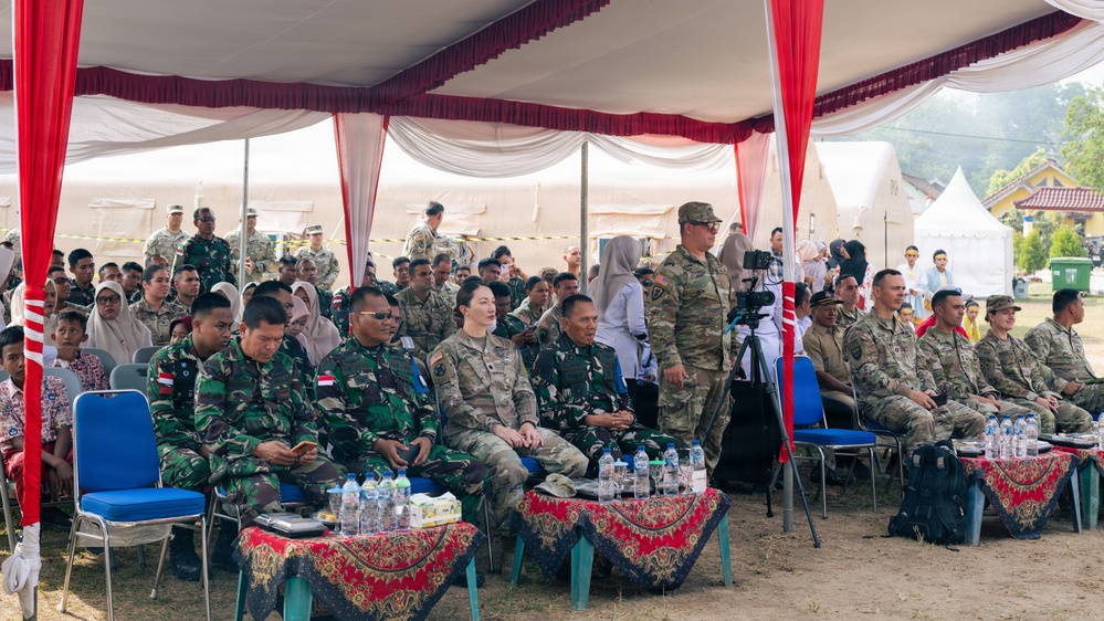 A formal military gathering is taking place under a large white and red tent, featuring personnel from what appears to be two different armed forces seated together. Indonesian military personnel in green camouflage uniforms are seated alongside soldiers in MultiCam/OCP uniforms consistent with U.S. Army dress. The setting suggests a joint military ceremony or exercise event, with additional tents and personnel visible in the background.
