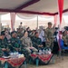 A formal military gathering is taking place under a large white and red tent, featuring personnel from what appears to be two different armed forces seated together. Indonesian military personnel in green camouflage uniforms are seated alongside soldiers in MultiCam/OCP uniforms consistent with U.S. Army dress. The setting suggests a joint military ceremony or exercise event, with additional tents and personnel visible in the background.