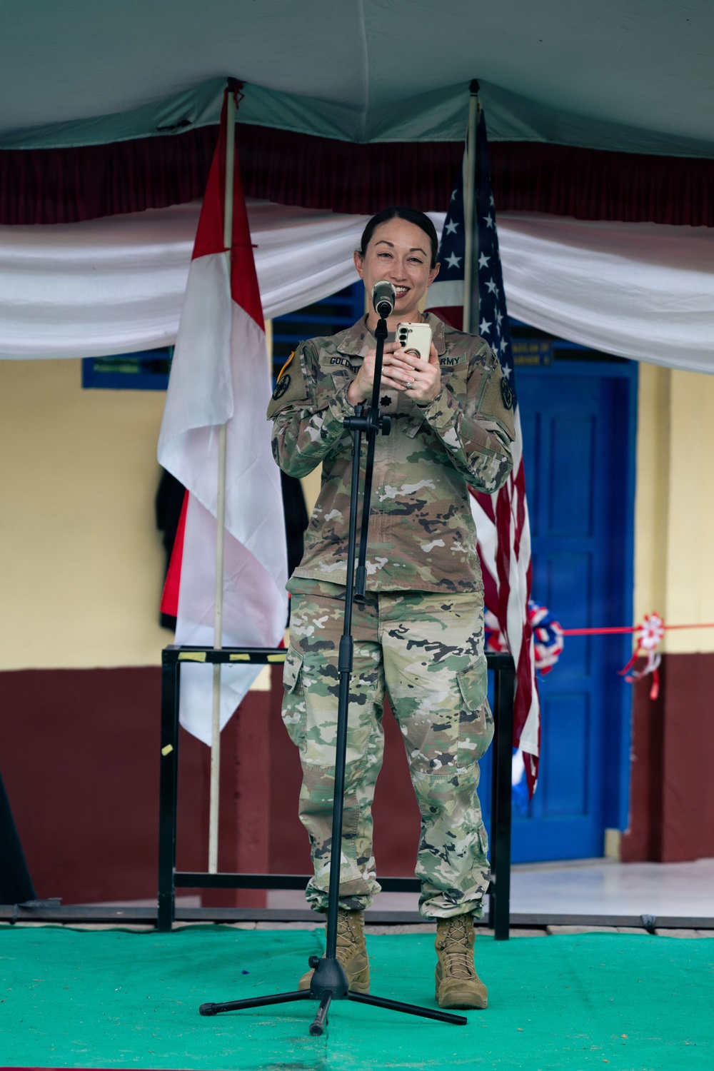 A female U.S. Army soldier in MultiCam (OCP) uniform stands at a microphone on a stage, holding a smartphone and smiling. She appears to be speaking or presenting at a joint military ceremony, flanked by the Indonesian flag (red and white) and the U.S. flag. The setting suggests a bilateral military engagement or cooperation event.