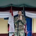 A female U.S. Army soldier in MultiCam (OCP) uniform stands at a microphone on a stage, holding a smartphone and smiling. She appears to be speaking or presenting at a joint military ceremony, flanked by the Indonesian flag (red and white) and the U.S. flag. The setting suggests a bilateral military engagement or cooperation event.
