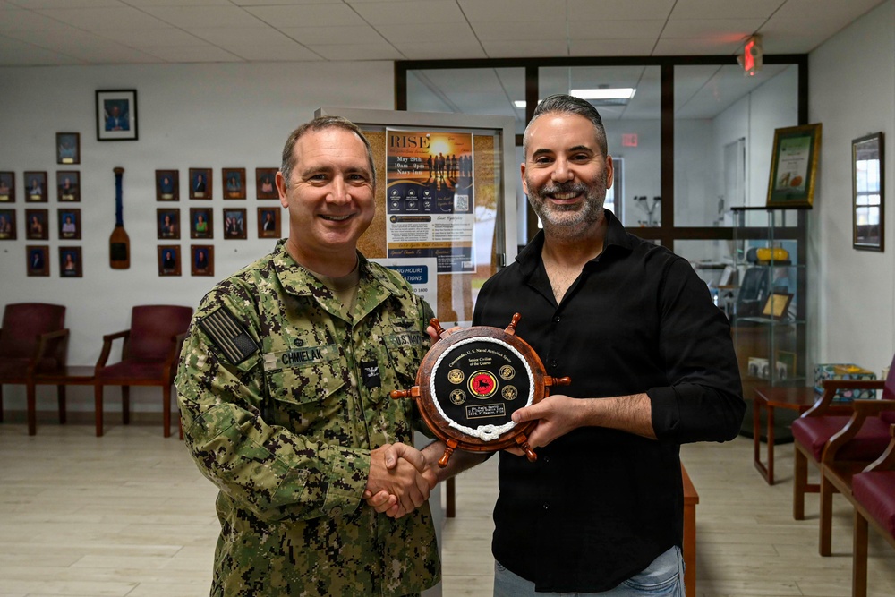 A U.S. Navy servicemember in camouflage uniform is shaking hands with a civilian man while both hold a ship's wheel-shaped award plaque. The setting appears to be a military administrative or community office, with portrait photos on the wall and a 'RISE' event poster visible in the background. This appears to be a formal recognition ceremony presenting a 'Senior Civilian of the Quarter' award.