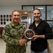A U.S. Navy servicemember in camouflage uniform is shaking hands with a civilian man while both hold a ship's wheel-shaped award plaque. The setting appears to be a military administrative or community office, with portrait photos on the wall and a 'RISE' event poster visible in the background. This appears to be a formal recognition ceremony presenting a 'Senior Civilian of the Quarter' award.