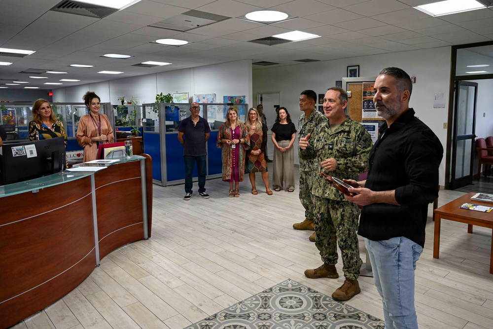 A group of approximately 10-12 people are gathered in what appears to be a military administrative office or welcome center. Two individuals in U.S. Navy camouflage uniforms stand alongside a civilian man in a black shirt who appears to be addressing the group. The setting suggests an official visit, briefing, or recognition ceremony.