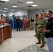 A group of approximately 10-12 people are gathered in what appears to be a military administrative office or welcome center. Two individuals in U.S. Navy camouflage uniforms stand alongside a civilian man in a black shirt who appears to be addressing the group. The setting suggests an official visit, briefing, or recognition ceremony.