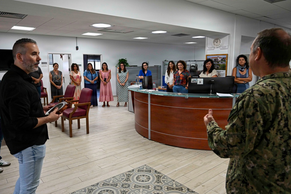 A group of approximately ten women are gathered in what appears to be an office reception area, listening to two men who appear to be presenting or speaking. One man is in civilian clothing holding a framed item, while the other is wearing military camouflage fatigues. The setting suggests an official briefing, orientation, or presentation event.