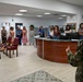 A group of approximately ten women are gathered in what appears to be an office reception area, listening to two men who appear to be presenting or speaking. One man is in civilian clothing holding a framed item, while the other is wearing military camouflage fatigues. The setting suggests an official briefing, orientation, or presentation event.