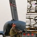 A military service member in camouflage uniform is working on a flight line, securing or releasing cargo straps on a Kaufman Trailer loaded with palletized cargo. A large military transport aircraft (appears to be a C-17 or similar) is visible in the background. The scene depicts a logistics/airlift operation, likely at a military airfield.