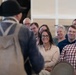 A historical reenactor dressed in 18th-century colonial attire, including a tricorn hat and carrying a musket, is presenting to a seated audience in what appears to be a historic building or meeting hall. The audience members are visibly engaged and smiling, suggesting an entertaining and informative presentation. The setting and costume indicate this is likely a living history demonstration or guided tour at a colonial-era historic site.