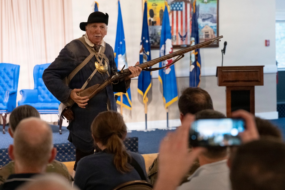 A historical reenactor dressed in 18th-century colonial American attire is presenting to an audience in what appears to be a military facility. He is holding a period-accurate flintlock musket and wearing a tricorn hat, cross-belt, and colonial-era clothing. Several audience members are visible from behind, with one person recording the presentation on a smartphone.