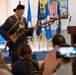 A historical reenactor dressed in 18th-century colonial American attire is presenting to an audience in what appears to be a military facility. He is holding a period-accurate flintlock musket and wearing a tricorn hat, cross-belt, and colonial-era clothing. Several audience members are visible from behind, with one person recording the presentation on a smartphone.