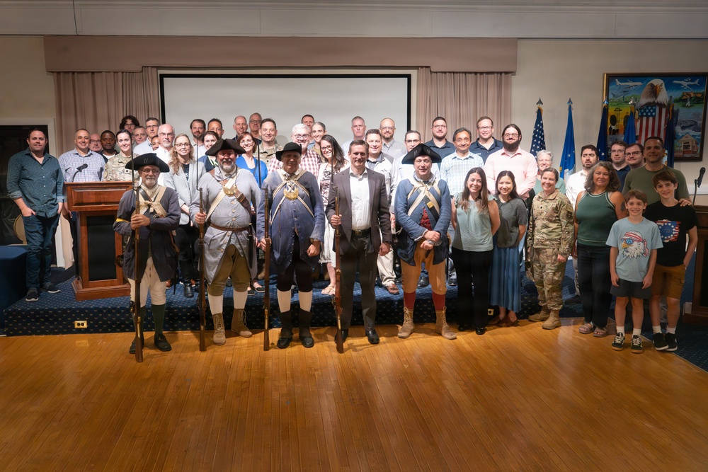 A large group photo taken in what appears to be a formal military or government venue, featuring approximately 40-50 people including several individuals dressed in American Revolutionary War-era colonial military uniforms holding muskets. The event appears to be a formal ceremony or commemorative gathering, evidenced by the presence of U.S. Air Force flags, a podium, and a projection screen in the background. One person in modern military camouflage uniform (ACU) is also visible among the civilian attendees.
