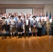 A large group photo taken in what appears to be a formal military or government venue, featuring approximately 40-50 people including several individuals dressed in American Revolutionary War-era colonial military uniforms holding muskets. The event appears to be a formal ceremony or commemorative gathering, evidenced by the presence of U.S. Air Force flags, a podium, and a projection screen in the background. One person in modern military camouflage uniform (ACU) is also visible among the civilian attendees.