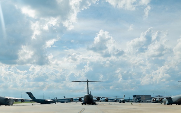 Multiple large military transport aircraft are parked on a wide concrete flight line apron at a military air base. The aircraft are arranged in a symmetrical formation facing away from the camera, with a yellow centerline stripe visible on the tarmac. The scene is photographed under a partly cloudy sky with bright sunlight breaking through.