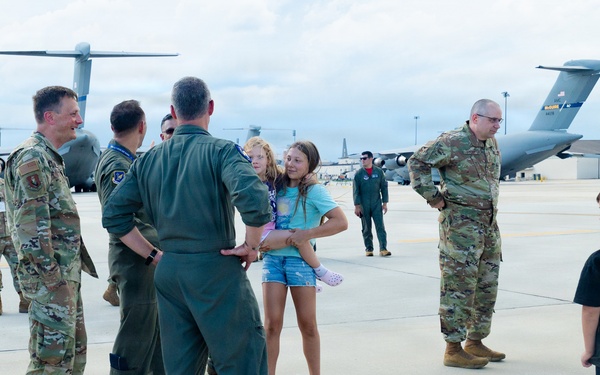 A military homecoming or greeting ceremony on an airfield tarmac, where uniformed personnel in OCP (Operational Camouflage Pattern) and flight suits are interacting with civilians, including children. A pilot in a green flight suit is holding or embracing a young girl, while a boy in a black shirt and blue Crocs stands nearby. Large military transport aircraft, consistent with C-17 Globemaster IIIs, are visible in the background.