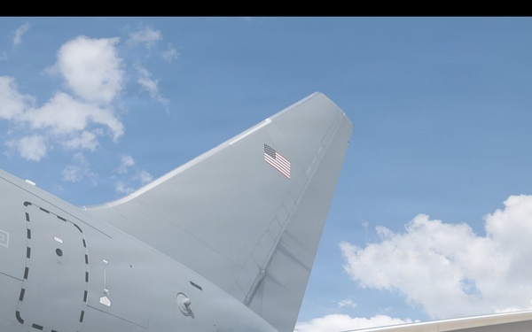 The image shows the tail section and horizontal stabilizer of a U.S. military aircraft, photographed from a low angle looking upward. The aircraft is painted in a light gray military finish and bears a U.S. Air Force roundel insignia on the fuselage and an American flag on the vertical stabilizer. The photo is taken outdoors under a partly cloudy blue sky.