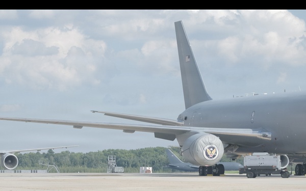 Two large gray military aircraft are parked on a concrete flight line apron, with a large hangar visible on the left side. The aircraft appear to be KC-46 Pegasus tankers based on their distinctive wide-body configuration and gray military paint scheme. Ground support equipment and personnel are visible near the foreground aircraft.