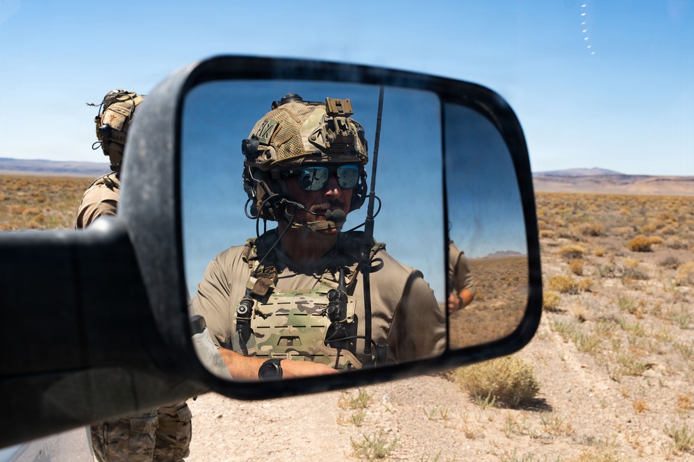 A military or special operations personnel is reflected in a vehicle's side mirror in an arid desert environment. The soldier is wearing full tactical gear including a combat helmet, plate carrier vest, communications headset, and sunglasses. A second soldier is partially visible in the background outside the mirror's reflection.