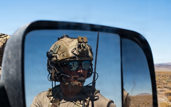 A military or special operations personnel is reflected in a vehicle's side mirror in an arid desert environment. The soldier is wearing full tactical gear including a combat helmet, plate carrier vest, communications headset, and sunglasses. A second soldier is partially visible in the background outside the mirror's reflection.