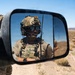 A military or special operations personnel is reflected in a vehicle's side mirror in an arid desert environment. The soldier is wearing full tactical gear including a combat helmet, plate carrier vest, communications headset, and sunglasses. A second soldier is partially visible in the background outside the mirror's reflection.