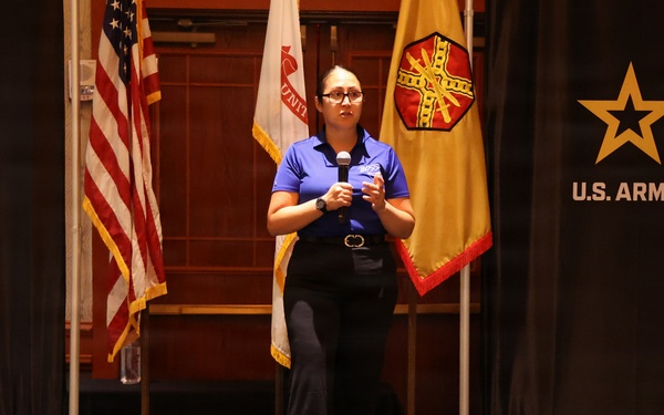 A woman in a blue polo shirt stands on a stage speaking into a microphone at a U.S. Army BOSS (Better Opportunities for Single Soldiers) event. She is positioned in front of multiple flags including the American flag and a U.S. Army flag, with branded backdrops on either side. On the stage floor in front of her are a framed award/plaque and a small bouquet of flowers.