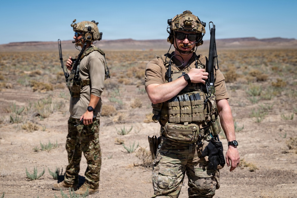 Two armed military or special operations personnel are standing in an arid desert environment with sparse vegetation. Both individuals are equipped with tactical gear including helmets, plate carriers, weapons, and communication equipment. The setting appears to be a training exercise or operational deployment in a desert region.